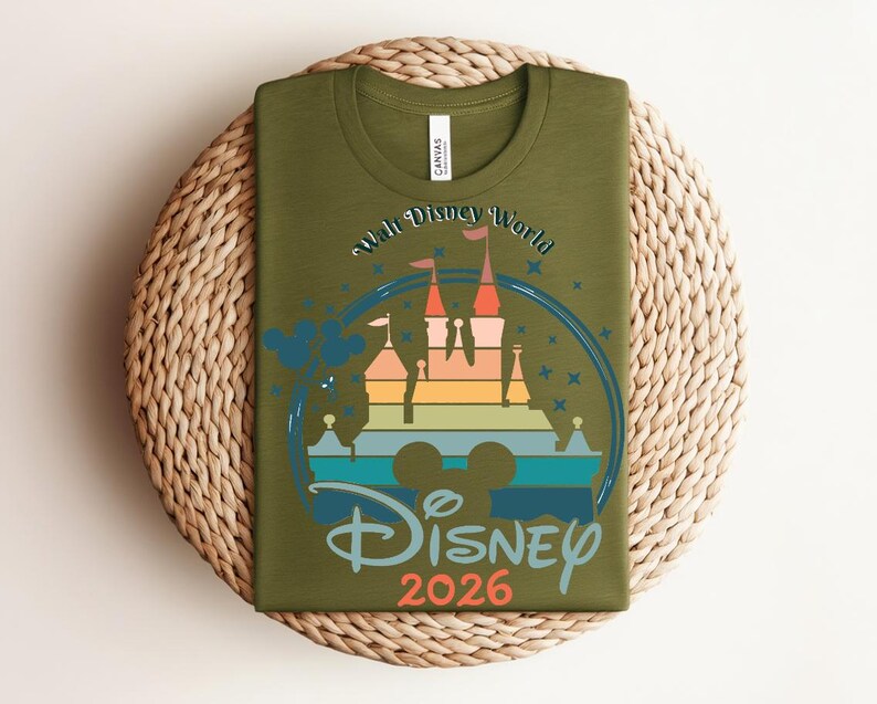 Op de afbeelding: Olijfgroen T-shirt met een kleurrijke afbeelding van een kasteel, Mickey Mouse oren en de woorden "Walt Disney World" en "Disney 2026". Het ontwerp bevat blauwe, oranje, gele en groene elementen.