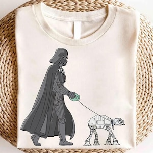 May include: Cream-colored t-shirt featuring a cartoon illustration of Darth Vader walking an AT-AT walker on a leash. Vader is depicted in his signature black helmet and cape. The AT-AT is drawn in a detailed, mechanical style.