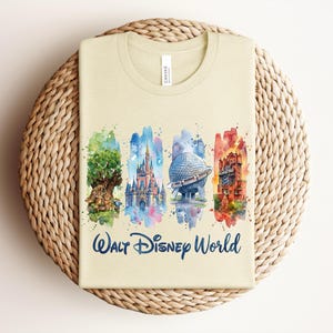 May include: A light beige t-shirt featuring watercolor illustrations of iconic Walt Disney World landmarks. The design includes the Tree of Life, Cinderella Castle, Spaceship Earth, and the Tower of Terror, with "Walt Disney World" text.