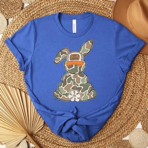 May include: A royal blue t-shirt featuring a cartoon bunny in camouflage print, wearing an orange hat with the text "Life is Good". The bunny has a white flower on its chest. The shirt is knotted at the bottom.