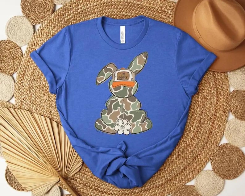 May include: A royal blue t-shirt featuring a cartoon bunny in camouflage print, wearing an orange hat with the text "Life is Good". The bunny has a white flower on its chest. The shirt is knotted at the bottom.