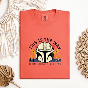 May include: Coral-coloured t-shirt with a Mandalorian helmet graphic and the text "THIS IS THE WAY." The design includes the words "BOUNTY HUNTER" and "CLAN OF TWO." The shirt is folded and displayed on a white surface with decorative elements.