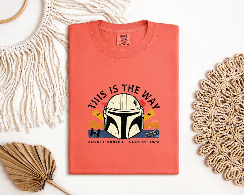 May include: Coral-coloured t-shirt with a Mandalorian helmet graphic and the text "THIS IS THE WAY." The design includes the words "BOUNTY HUNTER" and "CLAN OF TWO." The shirt is folded and displayed on a white surface with decorative elements.