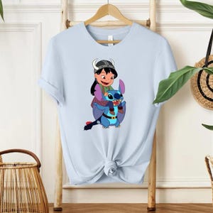 Może przedstawiać: Jasnoniebieska koszulka z kresk&oacute;wkową ilustracją dziewczynki jadącej na niebieskim stworze. Dziewczynka ma na sobie hełm i fioletowe ubrania. Stw&oacute;r ma duże uszy i pasek. Koszulka wisi na drewnianym wieszaku.