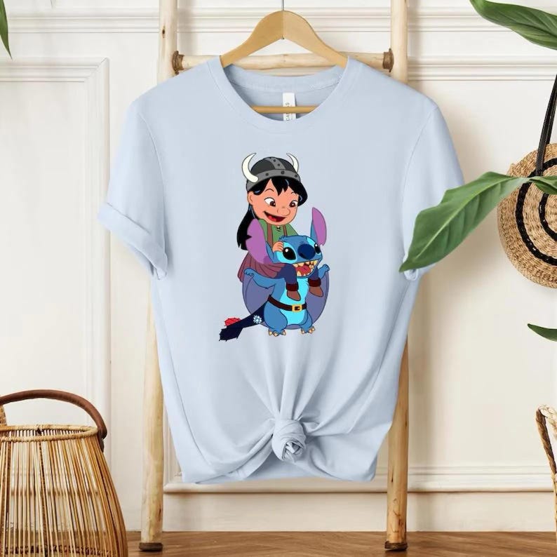 Może przedstawiać: Jasnoniebieska koszulka z kresk&oacute;wkową ilustracją dziewczynki jadącej na niebieskim stworze. Dziewczynka ma na sobie hełm i fioletowe ubrania. Stw&oacute;r ma duże uszy i pasek. Koszulka wisi na drewnianym wieszaku.