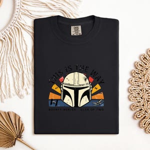 May include: Black t-shirt with a Mandalorian helmet graphic and the phrase "THIS IS THE WAY". The design incorporates a sunset-style backdrop in orange, yellow, and blue, along with the words "BOUNTY HUNTER CLAN OF TWO".