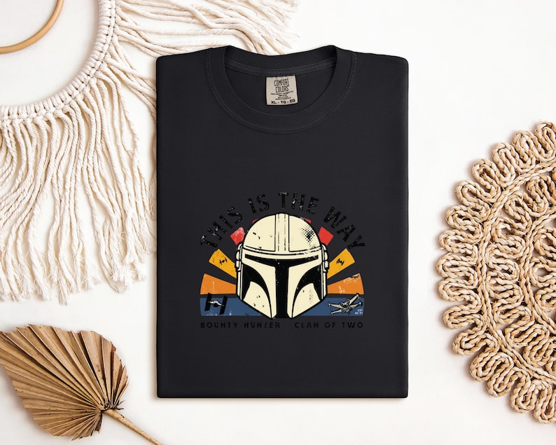 May include: Black t-shirt with a Mandalorian helmet graphic and the phrase "THIS IS THE WAY". The design incorporates a sunset-style backdrop in orange, yellow, and blue, along with the words "BOUNTY HUNTER CLAN OF TWO".
