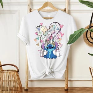 Pu&ograve; includere: T-shirt bianca con una grafica colorata del personaggio Stitch di Lilo & Stitch, con un castello e un design a orecchie di Topolino. Il design include colori pastello e stelle. La maglietta &egrave; appesa a una gruccia di legno.