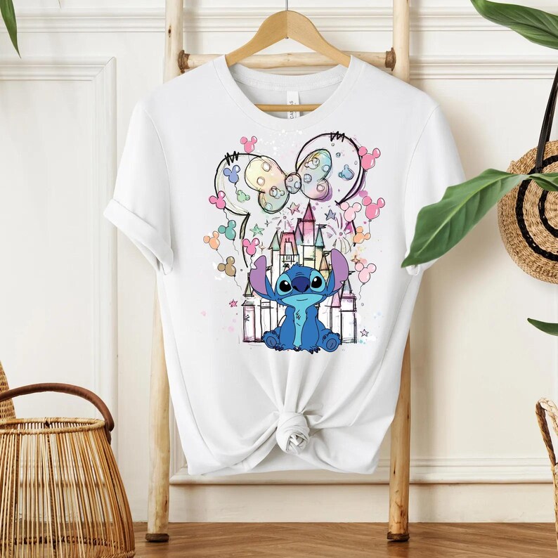 Pu&ograve; includere: T-shirt bianca con una grafica colorata del personaggio Stitch di Lilo & Stitch, con un castello e un design a orecchie di Topolino. Il design include colori pastello e stelle. La maglietta &egrave; appesa a una gruccia di legno.