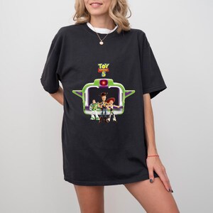 K&ouml;nnte beinhalten: Schwarzes T-Shirt mit einem Motiv aus dem Film Toy Story 5. Das Design zeigt Woody, Buzz Lightyear und Jessie in einem gr&uuml;n-wei&szlig;en Rahmen. Das T-Shirt hat kurze &Auml;rmel und einen Rundhalsausschnitt.