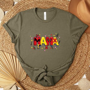 May include: Olive green t-shirt with the word "MAMA" in red and yellow, featuring cartoon characters and heart accents. The shirt is made of a soft material and has a crew neck.