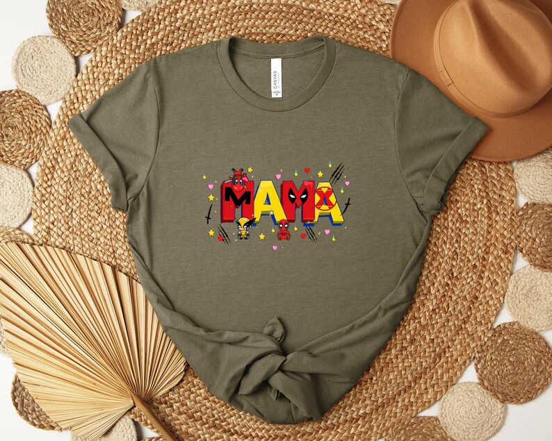 May include: Olive green t-shirt with the word "MAMA" in red and yellow, featuring cartoon characters and heart accents. The shirt is made of a soft material and has a crew neck.