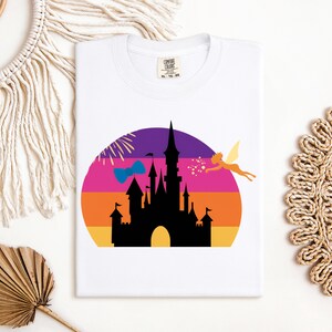 May include: White t-shirt featuring a colorful graphic of a castle silhouette against a sunset backdrop. A fairy and a bow are also included in the design. The shirt has a tag that says "Comfort Colors".