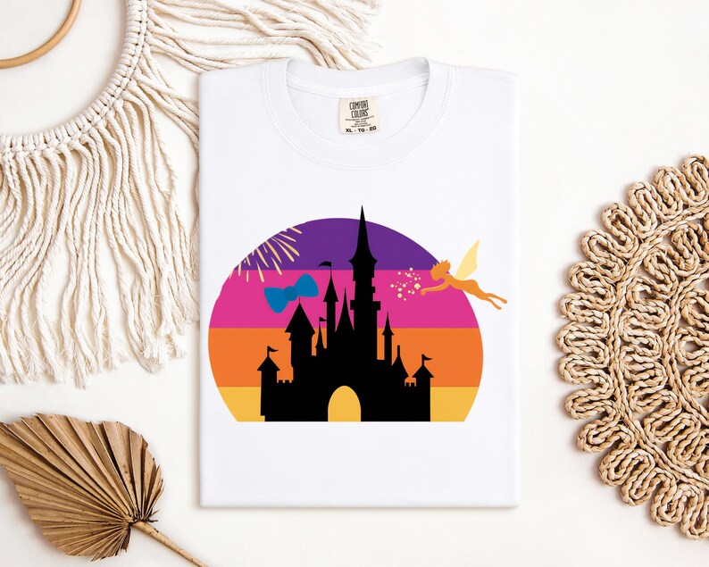 May include: White t-shirt featuring a colorful graphic of a castle silhouette against a sunset backdrop. A fairy and a bow are also included in the design. The shirt has a tag that says "Comfort Colors".