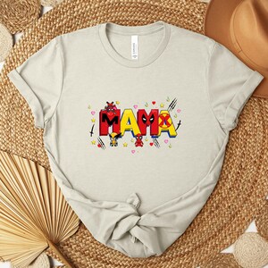 May include: A light beige t-shirt with the word "MAMA" in red and yellow block letters, decorated with cartoon characters and hearts. The shirt is made of a soft material and has short sleeves.