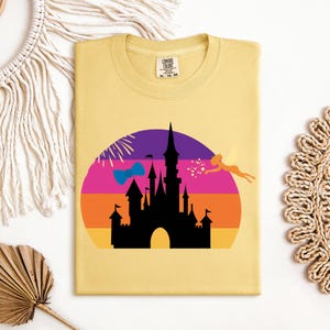 May include: A light yellow t-shirt featuring a colorful graphic of a castle silhouette against a sunset backdrop. The design includes a fairy, fireworks, and a bow. The shirt has a crew neck and a tag in the upper right corner.