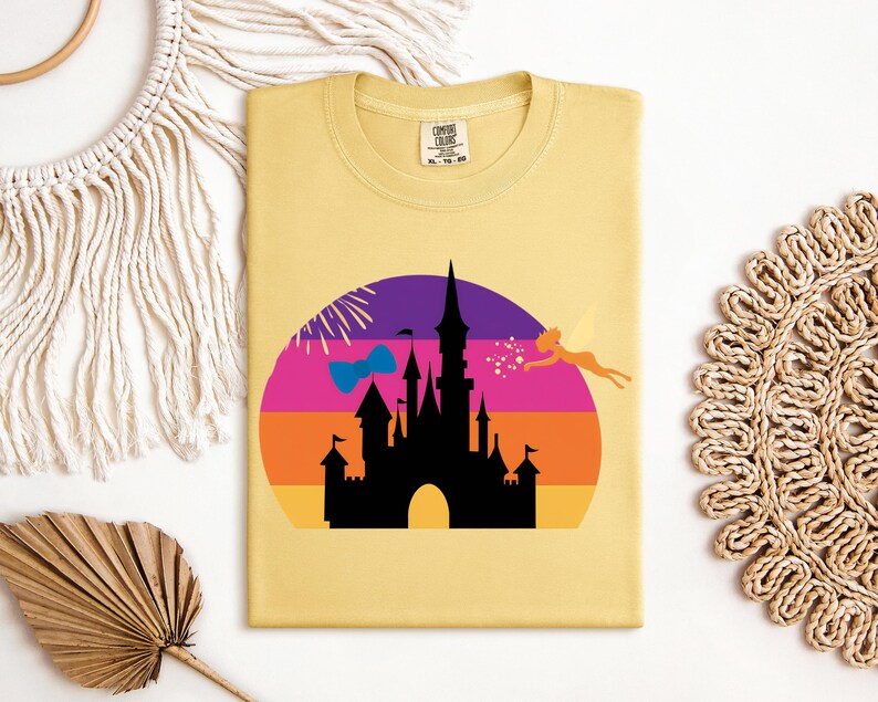 May include: A light yellow t-shirt featuring a colorful graphic of a castle silhouette against a sunset backdrop. The design includes a fairy, fireworks, and a bow. The shirt has a crew neck and a tag in the upper right corner.