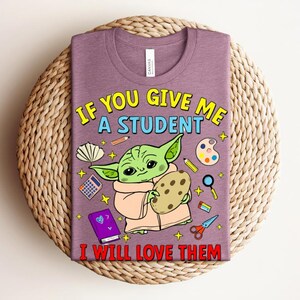 May include: A purple t-shirt featuring a cartoon image of a green alien-like character holding a cookie. The text on the shirt reads "IF YOU GIVE ME A STUDENT I WILL LOVE THEM" in yellow and red. The shirt is on a woven surface.