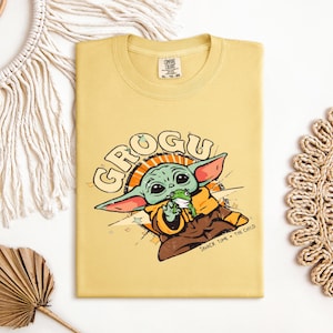 May include: A yellow t-shirt featuring a cartoon image of Grogu, also known as The Child, from the Star Wars series. The graphic includes the word "GROGU" in a retro font, and the phrase "SNACK TIME - THE CHILD".