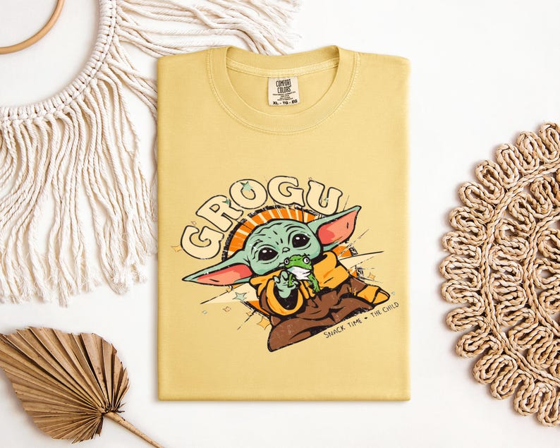 May include: A yellow t-shirt featuring a cartoon image of Grogu, also known as The Child, from the Star Wars series. The graphic includes the word "GROGU" in a retro font, and the phrase "SNACK TIME - THE CHILD".