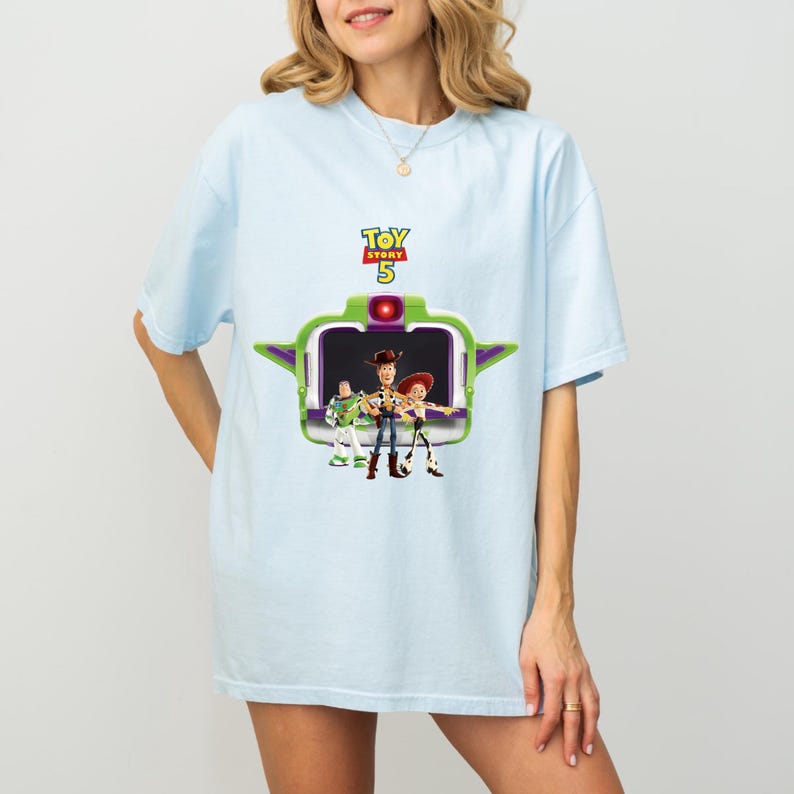 K&ouml;nnte beinhalten: Hellblaues T-Shirt mit einer Grafik von Charakteren aus Toy Story 5. Die Grafik zeigt Woody, Buzz Lightyear und Jessie, umrahmt von einem gr&uuml;nen und lila Design. Das Toy Story 5-Logo befindet sich oben.