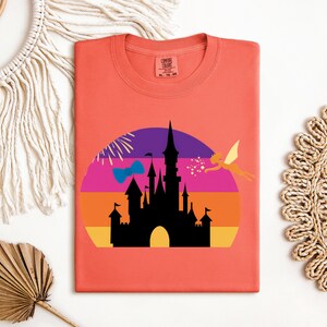 May include: Coral-colored t-shirt featuring a graphic of a black castle silhouette set against a colorful sunset design. A fairy is flying to the right. The shirt has a round neckline and short sleeves. The tag says "Comfort Colors".