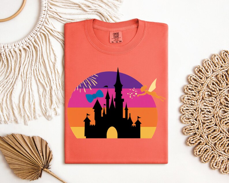 May include: Coral-colored t-shirt featuring a graphic of a black castle silhouette set against a colorful sunset design. A fairy is flying to the right. The shirt has a round neckline and short sleeves. The tag says "Comfort Colors".