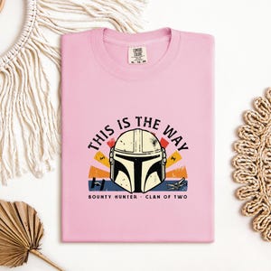May include: A pale pink t-shirt with a graphic of a Mandalorian helmet and the phrase "THIS IS THE WAY". The design includes a sunset-style background with the words "BOUNTY HUNTER" and "CLAN OF TWO".