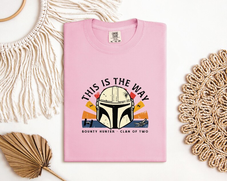 May include: A pale pink t-shirt with a graphic of a Mandalorian helmet and the phrase "THIS IS THE WAY". The design includes a sunset-style background with the words "BOUNTY HUNTER" and "CLAN OF TWO".