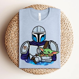 May include: Light blue t-shirt featuring a cartoon illustration of a Mandalorian character holding a green alien. The Mandalorian is wearing a helmet and armor. The alien is small with large ears. The shirt is folded on a woven surface.