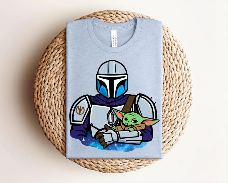 May include: Light blue t-shirt featuring a cartoon illustration of a Mandalorian character holding a green alien. The Mandalorian is wearing a helmet and armor. The alien is small with large ears. The shirt is folded on a woven surface.