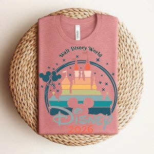 Op de afbeelding: Een oudroze t-shirt met een afbeelding van een kasteel in een regenboogkleurverloop, omringd door een blauwe cirkel en sterren. De tekst "Walt Disney World" en "Disney 2026" zijn ook zichtbaar. Het shirt is opgevouwen op een geweven, ronde placemat.