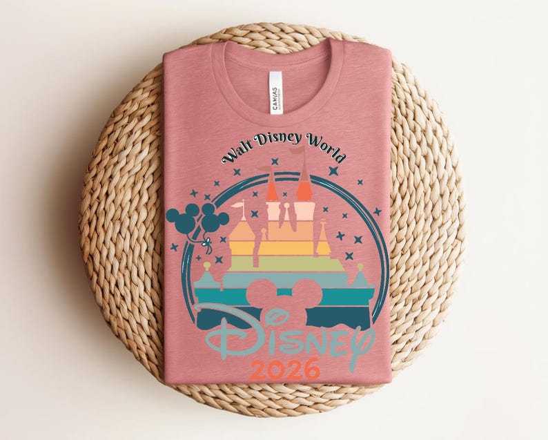 Op de afbeelding: Een oudroze t-shirt met een afbeelding van een kasteel in een regenboogkleurverloop, omringd door een blauwe cirkel en sterren. De tekst "Walt Disney World" en "Disney 2026" zijn ook zichtbaar. Het shirt is opgevouwen op een geweven, ronde placemat.