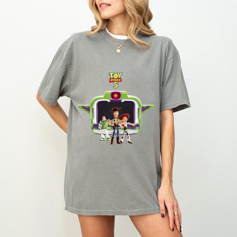 K&ouml;nnte beinhalten: Graues T-Shirt mit einem Toy Story 5-Motiv. Das Design zeigt Buzz Lightyear, Woody und Jessie in einem gr&uuml;n-wei&szlig;en Rahmen. Der Text "TOY STORY 5" befindet sich in Gelb und Gr&uuml;n &uuml;ber dem Rahmen.