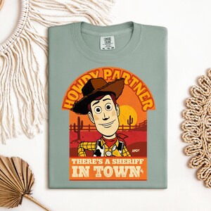 K&ouml;nnte beinhalten: Salbeigr&uuml;nes T-Shirt mit einer Grafik von Woody aus Toy Story. Das Design enth&auml;lt den Text "HOWDY PARTNER" &uuml;ber Woodys Bild und "THERE'S A SHERIFF IN TOWN" darunter. Die Grafik ist vor einer W&uuml;sten-Sonnenuntergangs-Kulisse platziert.