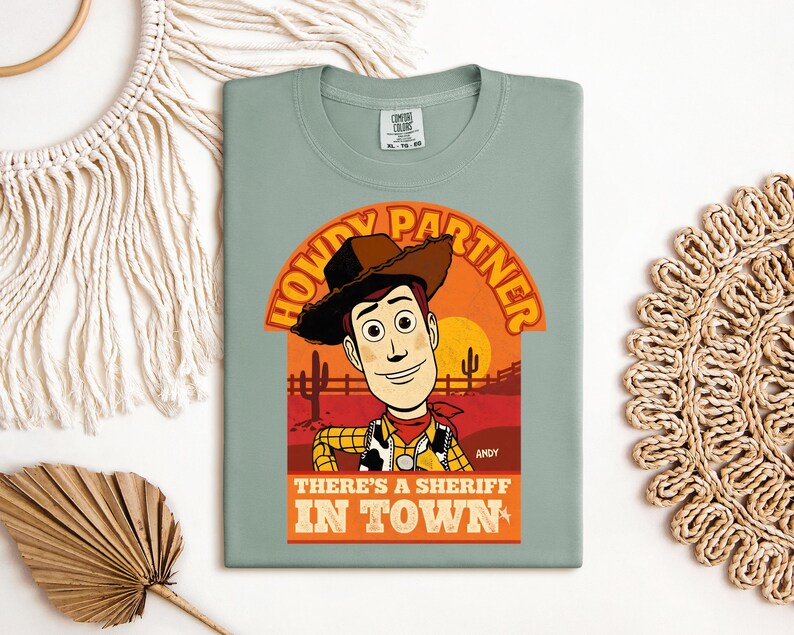 K&ouml;nnte beinhalten: Salbeigr&uuml;nes T-Shirt mit einer Grafik von Woody aus Toy Story. Das Design enth&auml;lt den Text "HOWDY PARTNER" &uuml;ber Woodys Bild und "THERE'S A SHERIFF IN TOWN" darunter. Die Grafik ist vor einer W&uuml;sten-Sonnenuntergangs-Kulisse platziert.
