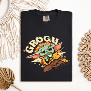 May include: Black t-shirt featuring a Grogu graphic. The design includes Grogu, a green alien character, with a frog in his mouth, surrounded by the word "GROGU" in a stylized font. The shirt is a Comfort Colors brand.