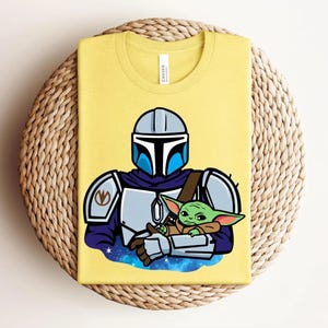 May include: Yellow t-shirt featuring a cartoon illustration of a Mandalorian character holding a green alien. The Mandalorian is wearing a blue and silver helmet and armor. The alien is green with large ears. The shirt is folded on a woven surface.