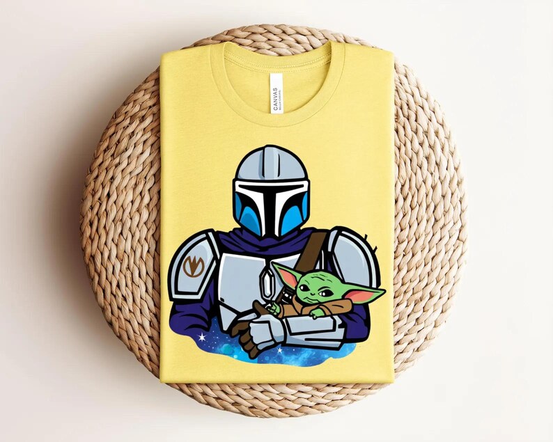 May include: Yellow t-shirt featuring a cartoon illustration of a Mandalorian character holding a green alien. The Mandalorian is wearing a blue and silver helmet and armor. The alien is green with large ears. The shirt is folded on a woven surface.