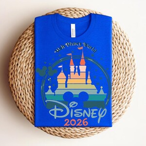 Op de afbeelding: Koningsblauw T-shirt met een kleurrijke afbeelding van een kasteel, de tekst "Walt Disney World" en "Disney 2026". Het ontwerp bevat Mickey Mouse oren en sterren. Het shirt ligt op een geweven, ronde placemat.