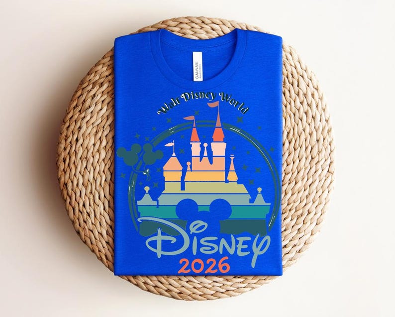 Op de afbeelding: Koningsblauw T-shirt met een kleurrijke afbeelding van een kasteel, de tekst "Walt Disney World" en "Disney 2026". Het ontwerp bevat Mickey Mouse oren en sterren. Het shirt ligt op een geweven, ronde placemat.