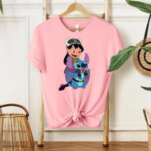 Może przedstawiać: Jasnor&oacute;żowa koszulka z kresk&oacute;wkową ilustracją dziewczynki w hełmie wikinga jadącej na niebieskim, obcym stworzeniu. Ilustracja wykorzystuje kolory niebieski, zielony, brązowy i czarny. Koszulka wisi na drewnianym wieszaku.