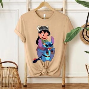 Może przedstawiać: Beżowa koszulka z kresk&oacute;wkową grafiką. Grafika przedstawia dziewczynkę w hełmie wikinga jadącą na niebieskim, obcym stworzeniu. Dziewczyna ma na sobie zielony i fioletowy str&oacute;j. Projekt jest kolorowy i szczeg&oacute;łowy.