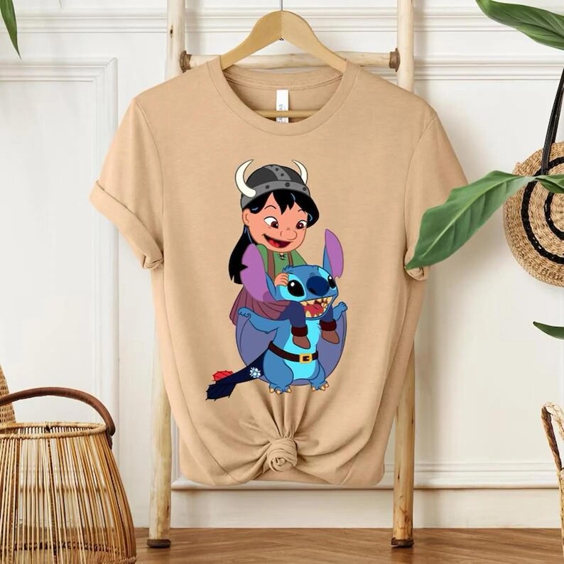 Może przedstawiać: Beżowa koszulka z kresk&oacute;wkową grafiką. Grafika przedstawia dziewczynkę w hełmie wikinga jadącą na niebieskim, obcym stworzeniu. Dziewczyna ma na sobie zielony i fioletowy str&oacute;j. Projekt jest kolorowy i szczeg&oacute;łowy.