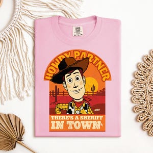K&ouml;nnte beinhalten: Hellrosa T-Shirt mit einer Grafik von Woody aus Toy Story. Die Grafik enth&auml;lt den Text "HOWDY PARTNER" und "THERE'S A SHERIFF IN TOWN". Das Design ist vor einer W&uuml;stenlandschaft mit Sonnenuntergang platziert.