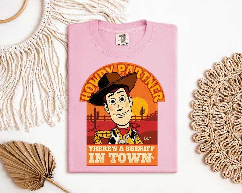 K&ouml;nnte beinhalten: Hellrosa T-Shirt mit einer Grafik von Woody aus Toy Story. Die Grafik enth&auml;lt den Text "HOWDY PARTNER" und "THERE'S A SHERIFF IN TOWN". Das Design ist vor einer W&uuml;stenlandschaft mit Sonnenuntergang platziert.