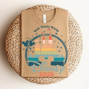 Op de afbeelding: Een bruin T-shirt met een kleurrijke afbeelding van een kasteel, Mickey Mouse-ballonnen en de tekst "Walt Disney World Disney 2026". Het ontwerp is omcirkeld door een blauwe ring met sterren.