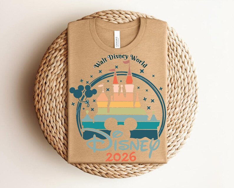 Op de afbeelding: Een bruin T-shirt met een kleurrijke afbeelding van een kasteel, Mickey Mouse-ballonnen en de tekst "Walt Disney World Disney 2026". Het ontwerp is omcirkeld door een blauwe ring met sterren.