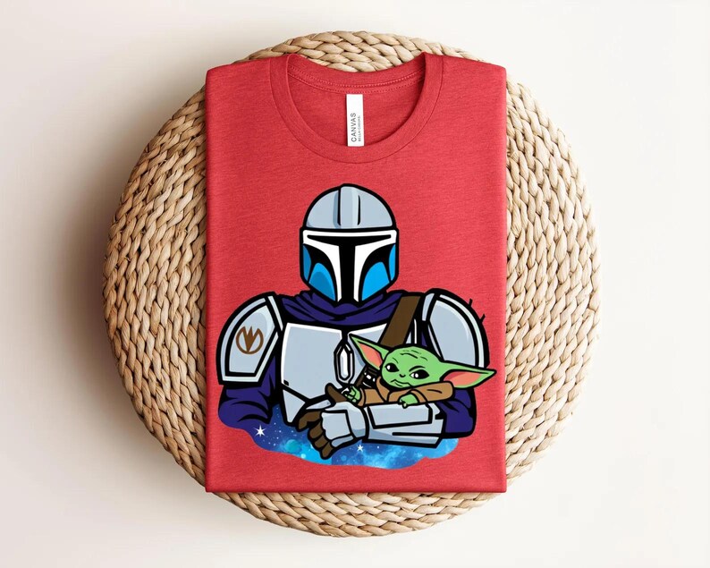 May include: Red t-shirt featuring a cartoon illustration of a character in a helmet holding a green alien. The character has a silver helmet with blue accents and is wearing silver armor. The alien is green with large ears. The shirt is folded on a woven surface.