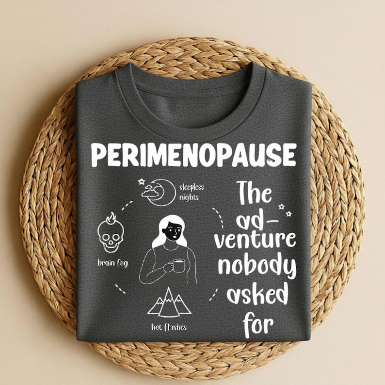 Puede incluir: Camiseta gris oscuro con la palabra "PERIMENOPAUSE" en blanco. La camiseta presenta ilustraciones de una calavera, una luna, una mujer y monta&ntilde;as, con texto que describe los s&iacute;ntomas. La frase "The ad-venture nobody asked for" tambi&eacute;n est&aacute; impresa.